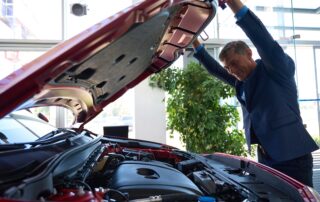 A middle-aged man in a blue suit looking under the open hood of a red car in a brightly lit showroom. He is smiling while inspecting the engine bay.
