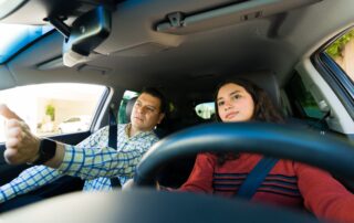 A young woman sitting in the driver’s seat of a car while a man in the passenger seat gestures forward, appearing to give her a driving lesson. Both are wearing seatbelts.