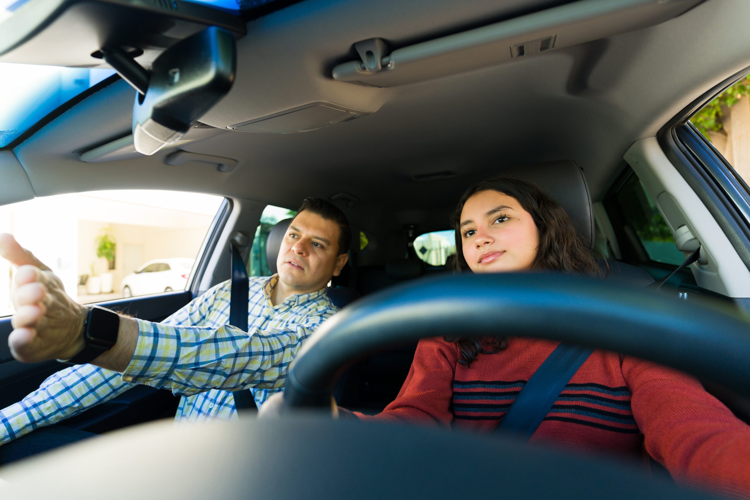 A young woman sitting in the driver’s seat of a car while a man in the passenger seat gestures forward, appearing to give her a driving lesson. Both are wearing seatbelts.
