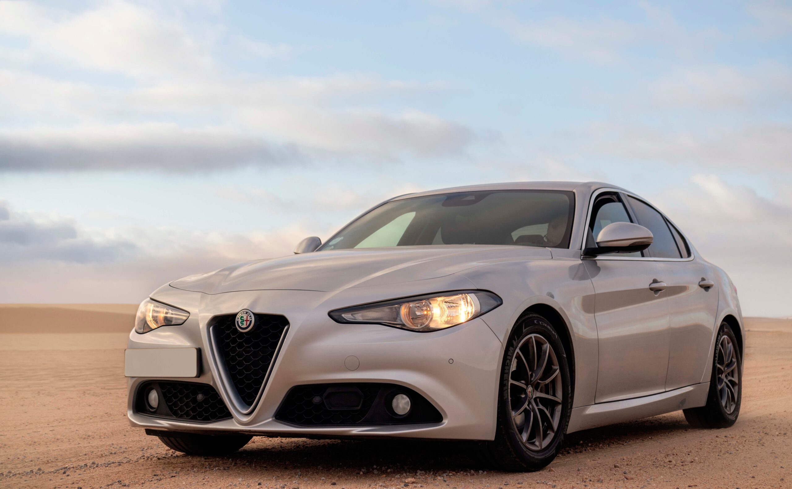 A silver Alfa Romeo Giulia sedan parked on a flat, sandy desert landscape under a pale blue sky filled with soft, scattered clouds. The car is angled toward the viewer, highlighting its distinctive triangular grille and illuminated headlights. The vast, empty sand dunes stretch out gently into the background.