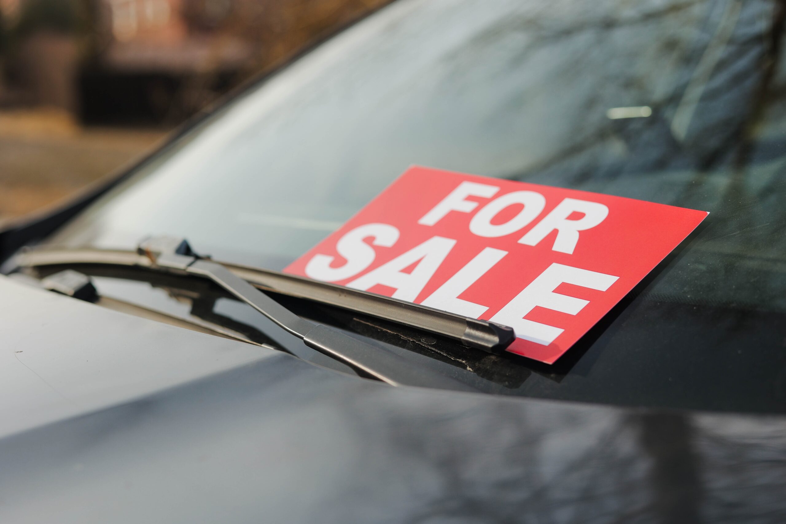 An alt text for this image could be: A close-up shot of a bright red sign with bold white lettering that reads "FOR SALE" resting on the lower portion of a black car's windshield. The windshield wiper is positioned just over the bottom edge of the sign. Reflections of bare tree branches are faintly visible on the glossy surface of the car's hood and windshield, with a blurred outdoor background.