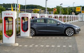 Side view of a grey Tesla Model 3 electric sedan parked in a designated stall at a Tesla Supercharging charging station lot.