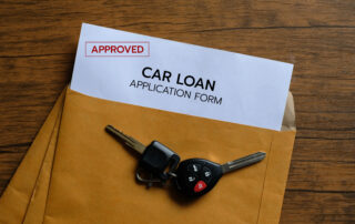 An approved car loan application form slipping out of a brown envelope next to car keys on a wooden table.