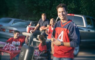 Smiling man in a football jersey holding tongs by a grill during an outdoor parking lot tailgate party with friends.