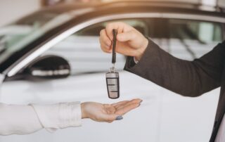 Car dealer handing over a key fob to a customer's open hand in front of a white vehicle.