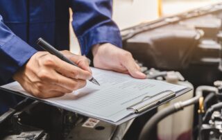 Auto mechanic filling out an inspection checklist on a clipboard over an open car engine bay.