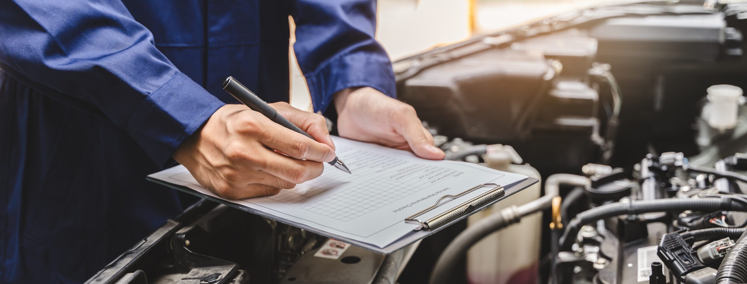 Auto mechanic filling out an inspection checklist on a clipboard over an open car engine bay.