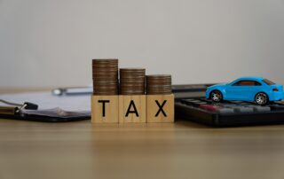 Alt Text: Car sales tax concept with TAX letter blocks, coin stacks, and a blue toy car on a calculator.