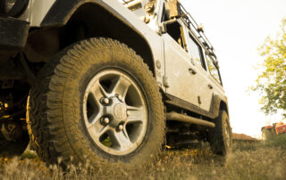 Low-angle close-up of a muddy off-road tire on a white Land Rover Defender parked in grassy terrain.